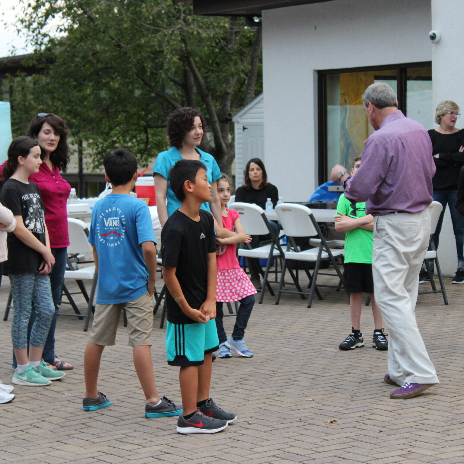 Group of children and adults gathered outdoors, listening to a man speaking.
